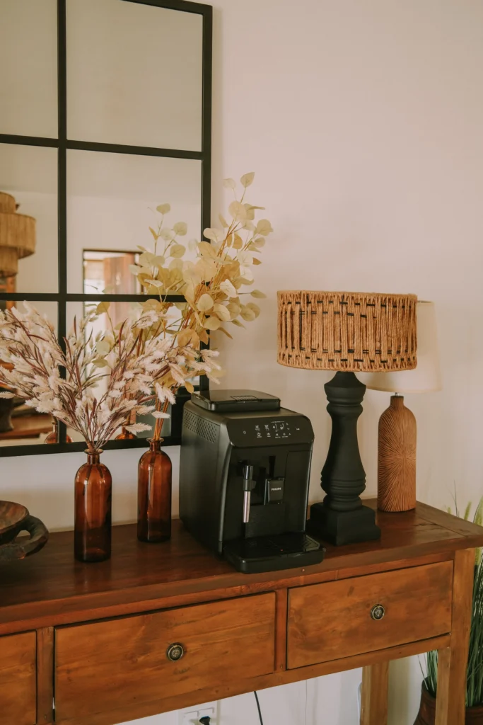 Rooikat safari themed interior with wooden sideboard, decorative dried stems in amber bottles, coffee machine, and woven lamp.