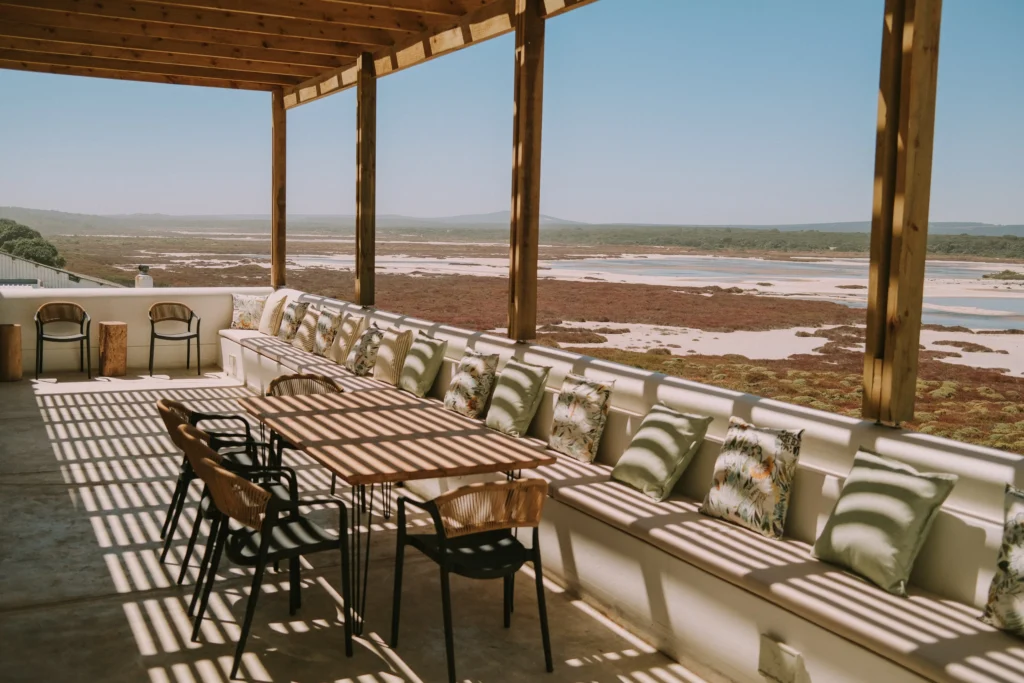 Rooikat safari coastal seating area with long couch and cushioned pillows under a shaded wooden pergola, overlooking a tidal flat and distant shoreline.