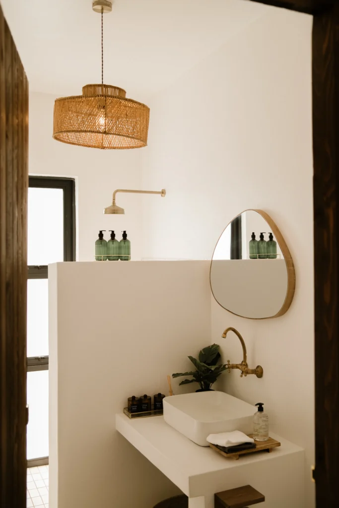 A stylish bathroom with a white sink, wall-mounted faucet, rounded mirror, wicker pendant light, and natural tones.